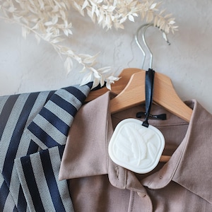Peut inclure: Une chemise boutonnée beige et un blazer à rayures bleu marine sont suspendus à un cintre en bois. Un désodorisant blanc de forme carrée avec un motif de plumes est suspendu au cintre. Des branches blanches décoratives sont en arrière-plan.