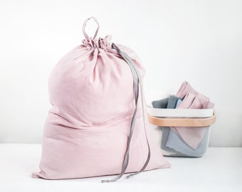 Puede incluir: Una bolsa de tela de color rosa claro con cierre de cordón, parte superior fruncida y una cinta gris larga y fina. Junto a la bolsa hay una pequeña cesta de plástico blanca que contiene varias piezas de tela dobladas en tonos rosa, gris y azul.