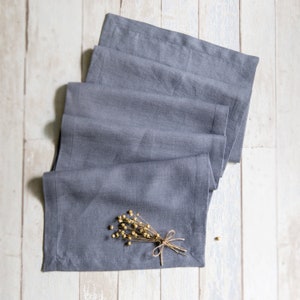 May include: A stack of gray linen napkins folded neatly on a white wooden surface. A small bouquet of dried flowers with a twine bow is placed on top of the napkins.