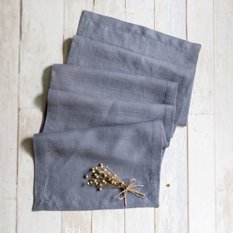 May include: A stack of gray linen napkins folded neatly on a white wooden surface. A small bouquet of dried flowers with a twine bow is placed on top of the napkins.