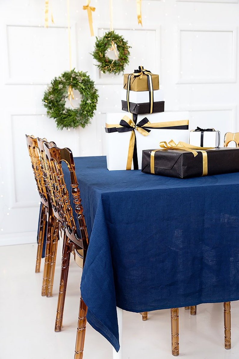 Puede incluir: Un mantel azul marino cubre una mesa con cuatro sillas de acr&iacute;lico transparente. Una pila de regalos envueltos con cinta negra y dorada se encuentra sobre la mesa. Dos coronas de laurel verde cuelgan de la pared detr&aacute;s de la mesa.