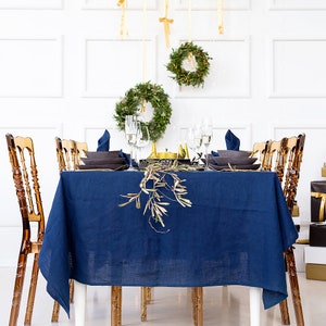 Puede incluir: Un mantel de lino azul preparado para una cena festiva con detalles dorados. La mesa est&aacute; puesta con platos blancos, vasos transparentes y cubiertos dorados. Hay ramas de olivo y vegetaci&oacute;n en la mesa.