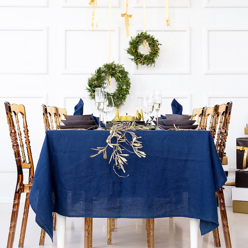 Puede incluir: Un mantel de lino azul preparado para una cena festiva con detalles dorados. La mesa est&aacute; puesta con platos blancos, vasos transparentes y cubiertos dorados. Hay ramas de olivo y vegetaci&oacute;n en la mesa.