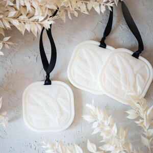 May include: Three square white wax tablets featuring a raised embossed leaf pattern on the front surface. Each tablet is suspended by a thin black ribbon loop attached to the top, designed for hanging as a decorative or aromatic ornament.