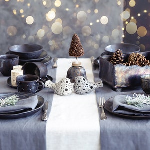 May include: A rustic table setting with black ceramic plates, mugs, and a small ceramic tree. The table is decorated with pine sprigs and a wooden box filled with pine cones.