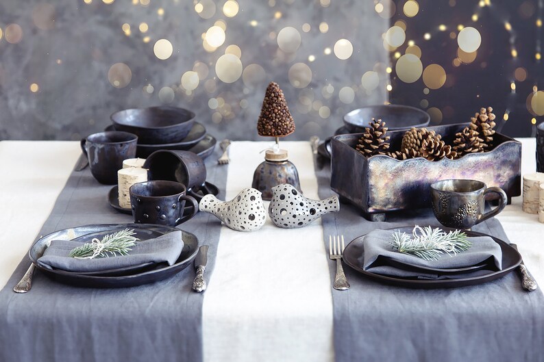 May include: A rustic table setting with black ceramic plates, mugs, and a small ceramic tree. The table is decorated with pine sprigs and a wooden box filled with pine cones.