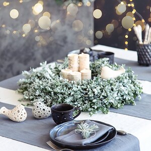 May include: A white tablecloth with a gray runner, a wreath of green leaves, and a place setting with a black mug, a gray napkin, and a black plate. There are two white bird figurines on the table.