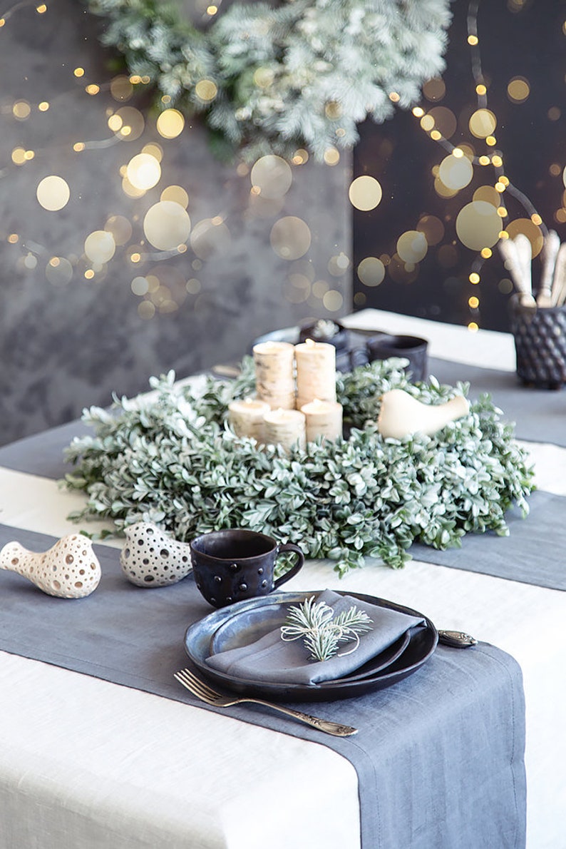 May include: A white tablecloth with a gray runner, a wreath of green leaves, and a place setting with a black mug, a gray napkin, and a black plate. There are two white bird figurines on the table.