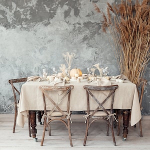 May include: A rectangular dining table covered with a beige linen tablecloth, surrounded by four dark wood cross-back chairs. The table is set with white plates, beige napkins, clear glassware, and a centerpiece of small white pumpkins and dried grasses.