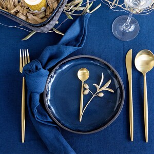 Puede incluir: Un plato de cena azul con una cuchara de oro y una rama de olivo sobre un mantel azul con cubiertos de oro y una servilleta azul.