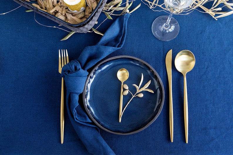 Puede incluir: Un plato de cena azul con una cuchara de oro y una rama de olivo sobre un mantel azul con cubiertos de oro y una servilleta azul.