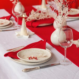 May include: A white table setting with a red runner, white plates, and a white napkin. A white candle holder with a lit candle is on the table. A glass of water and a wine glass are on the table. A vase with white flowers and red berries is on the table.