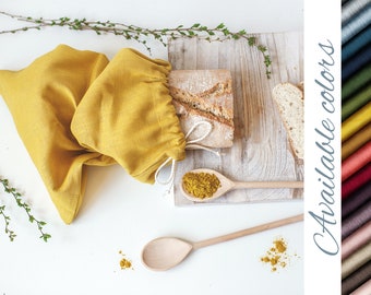 Pode incluir: Uma bolsa de pão de linho amarela com um pão dentro. A bolsa é amarrada com um cordão e há uma pequena tigela de especiarias ao lado. A imagem também mostra uma colher de madeira e uma tábua de cortar de madeira. O texto "Cores disponíveis" é mostrado no lado direito da imagem com uma variedade de amostras de tecido em diferentes cores.
