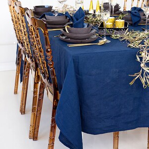 Puede incluir: Un mantel azul marino con cubiertos dorados y cuencos grises oscuros colocados en una mesa con sillas marrones. La mesa est&aacute; decorada con ramas de olivo y velas doradas.