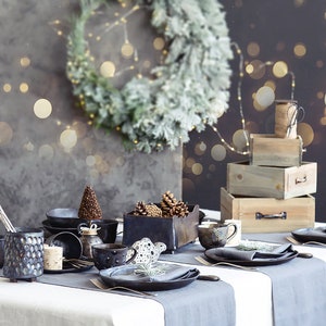 May include: A rustic table setting with a gray tablecloth and black ceramic plates, bowls, and mugs. The table is decorated with pine cones, a small Christmas tree, and a gray napkin with a sprig of greenery. The background features a wooden box stack and a wreath with lights.