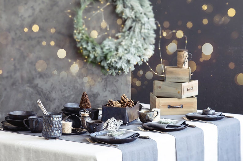 May include: A rustic table setting with a gray tablecloth and black ceramic plates, bowls, and mugs. The table is decorated with pine cones, a small Christmas tree, and a gray napkin with a sprig of greenery. The background features a wooden box stack and a wreath with lights.
