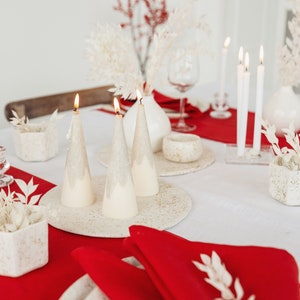 May include: A white tablecloth with a red runner and white ceramic candlesticks with lit candles. The candles are shaped like Christmas trees. There are white ceramic dishes and a white ceramic vase with dried white flowers. A red napkin with a white sprig of greenery is on a white plate.