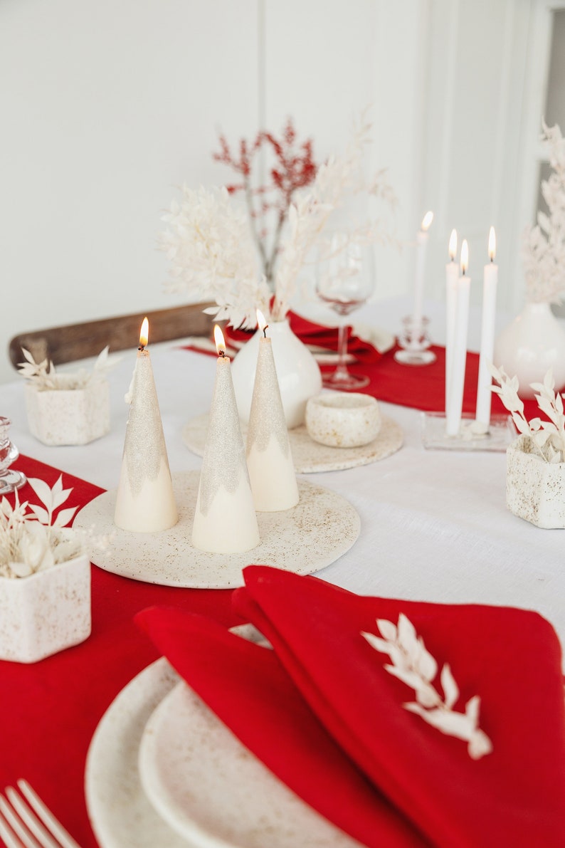 May include: A white tablecloth with a red runner and white ceramic candlesticks with lit candles. The candles are shaped like Christmas trees. There are white ceramic dishes and a white ceramic vase with dried white flowers. A red napkin with a white sprig of greenery is on a white plate.