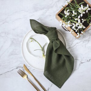 May include: A dark olive green linen napkin tied in a knot, placed on a white speckled ceramic plate. A single white snowdrop flower rests on the plate. Beside the plate are a gold-toned fork and knife, with a wooden planter of snowdrops nearby.