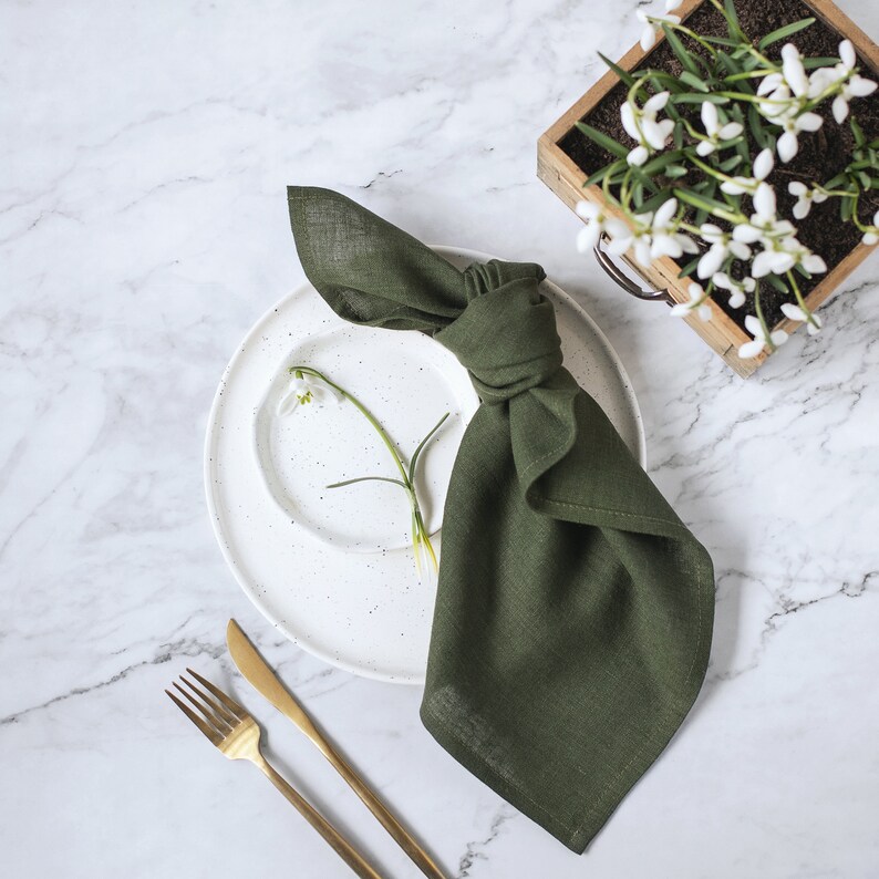 May include: A dark olive green linen napkin tied in a knot, placed on a white speckled ceramic plate. A single white snowdrop flower rests on the plate. Beside the plate are a gold-toned fork and knife, with a wooden planter of snowdrops nearby.