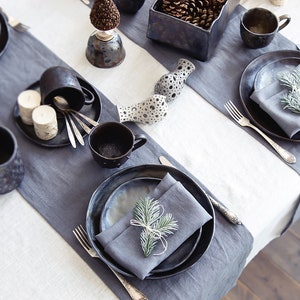 May include: A table setting with black and gray ceramic plates, bowls, and mugs. The table is set with gray napkins and silverware. There are small pine cones in a black ceramic bowl and a sprig of greenery on each plate.