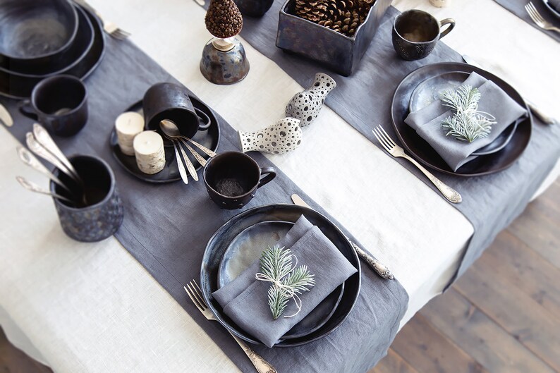 May include: A table setting with black and gray ceramic plates, bowls, and mugs. The table is set with gray napkins and silverware. There are small pine cones in a black ceramic bowl and a sprig of greenery on each plate.