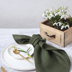 May include: A dark olive green linen napkin folded and placed on a white speckled ceramic plate. A small white plate holds a gold spoon and a single snowdrop flower. A wooden box filled with snowdrop flowers sits nearby with gold cutlery.