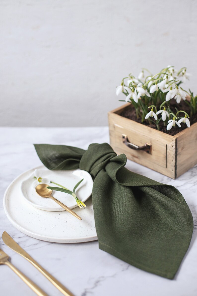 May include: A dark olive green linen napkin folded and placed on a white speckled ceramic plate. A small white plate holds a gold spoon and a single snowdrop flower. A wooden box filled with snowdrop flowers sits nearby with gold cutlery.