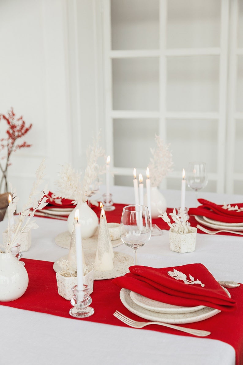 May include: A white table setting with red napkins, white plates, and white candles. The table is decorated with white flowers and greenery.
