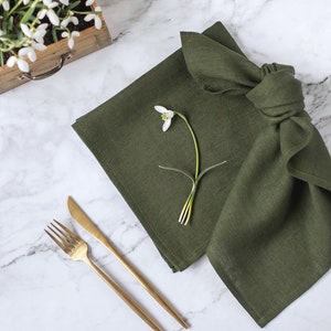 May include: A square olive green linen napkin folded with a knot, featuring a single white snowdrop flower placed on top. Beside the napkin are a gold-toned metal fork and a matching knife, all arranged on a white marble surface with grey veining.