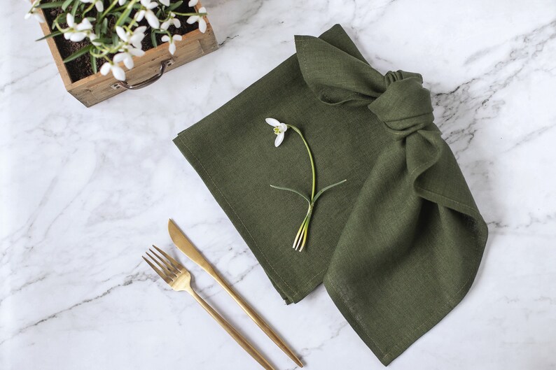 May include: A square olive green linen napkin folded with a knot, featuring a single white snowdrop flower placed on top. Beside the napkin are a gold-toned metal fork and a matching knife, all arranged on a white marble surface with grey veining.