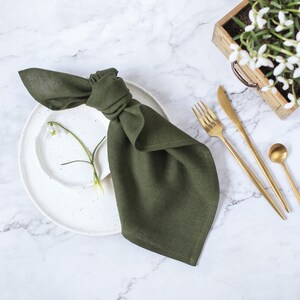 May include: A dark olive green linen napkin tied in a knot, resting on a white speckled ceramic plate. Beside the plate are gold-toned stainless steel flatware including a fork, knife, and small spoon, with a single white flower stem.