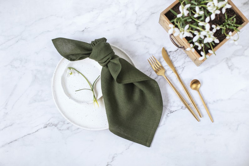 May include: A dark olive green linen napkin tied in a knot, resting on a white speckled ceramic plate. Beside the plate are gold-toned stainless steel flatware including a fork, knife, and small spoon, with a single white flower stem.