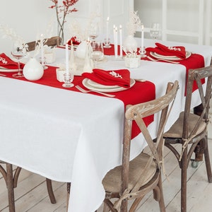 May include: A white tablecloth with red placemats and napkins set for a holiday dinner. The table is decorated with white candles and white vases with dried flowers. Two wooden chairs with woven seats are pulled up to the table.