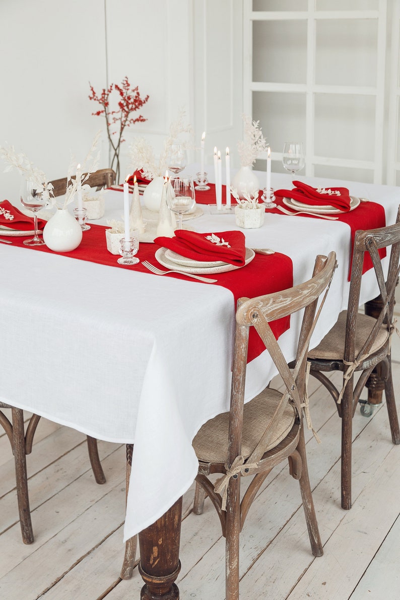May include: A white tablecloth with red placemats and napkins set for a holiday dinner. The table is decorated with white candles and white vases with dried flowers. Two wooden chairs with woven seats are pulled up to the table.