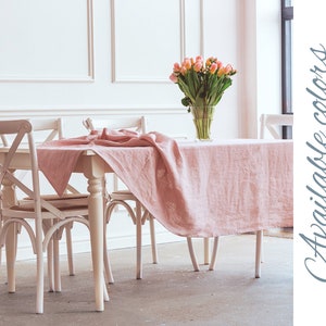 May include: A blush pink linen tablecloth draped over a white wooden table with four white chairs. The tablecloth is available in a variety of colors, including blue, green, yellow, orange, red, purple, brown, and gray.  Available colors.