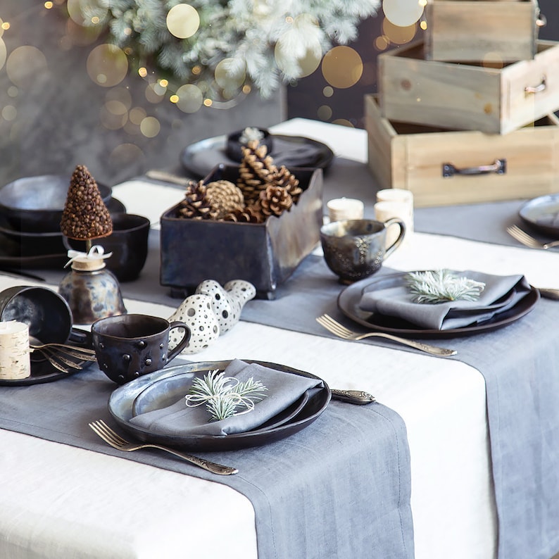 May include: A rustic table setting with a gray tablecloth and gray linen placemats. The table is set with black and gray ceramic dishes, a gray napkin, and a sprig of greenery. There are also small ceramic ornaments and a wooden box with pine cones.