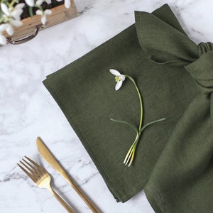 May include: A dark olive green linen napkin folded and placed next to a gold-toned metal fork and knife. A single white snowdrop flower with green accents rests on top of the fabric, which features a visible woven texture and hemmed edges.