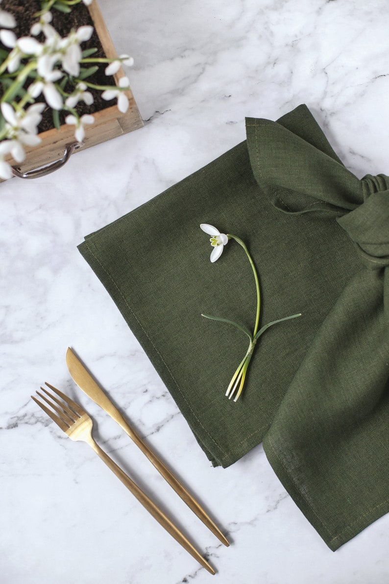May include: A dark olive green linen napkin folded and placed next to a gold-toned metal fork and knife. A single white snowdrop flower with green accents rests on top of the fabric, which features a visible woven texture and hemmed edges.