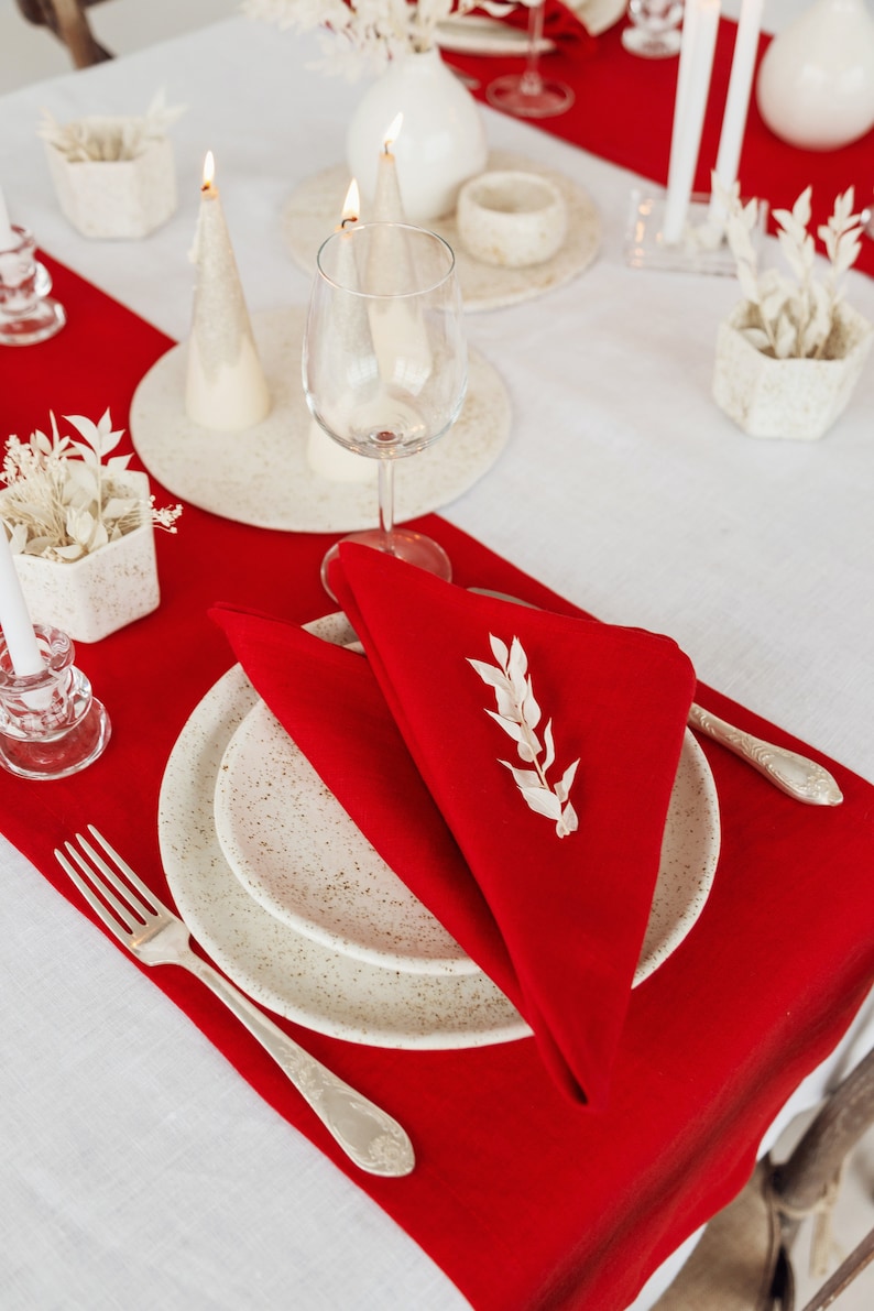 May include: A white table setting with a red runner. The setting includes a white plate, a white napkin folded into a triangle, a silver fork, and a sprig of dried white flowers. There are also two white candles and a glass of water.