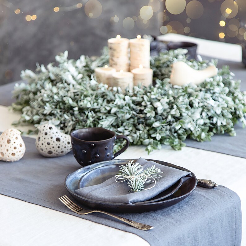May include: A rustic table setting with a gray linen tablecloth and a gray linen runner. A wreath of green leaves with white berries sits in the center of the table. A black ceramic mug, a white ceramic bird, and a gray ceramic bird are on the table. A black ceramic plate with a blue ceramic plate and a gray linen napkin are set for a place setting. A sprig of green leaves sits on top of the napkin.