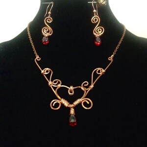 May include: A copper-colored wire jewelry set featuring a necklace and matching earrings. The necklace has a heart-shaped pendant with a teardrop-shaped red gemstone. The earrings have a spiral design with a similar red gemstone. The set is displayed on a black background.