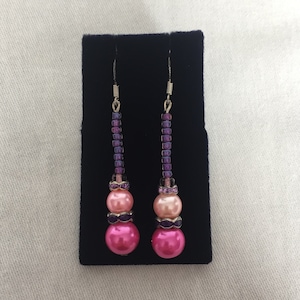May include: Dangle earrings with a combination of pink and purple beads. The earrings feature a large, round, hot pink bead at the bottom, followed by a smaller pink bead, and a series of purple beads. The earrings are displayed on a black surface.