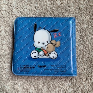 May include: A blue plastic wallet with a cartoon image of a white dog with black ears and a brown dog with a carrot. The wallet has the text "Pochacco" repeated on the front and back. The text "Sanrio Smiles" and "637112 450JXTOYO" is printed on the bottom of the wallet.