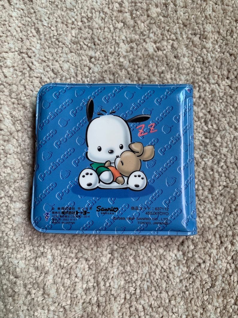 May include: A blue plastic wallet with a cartoon image of a white dog with black ears and a brown dog with a carrot. The wallet has the text "Pochacco" repeated on the front and back. The text "Sanrio Smiles" and "637112 450JXTOYO" is printed on the bottom of the wallet.