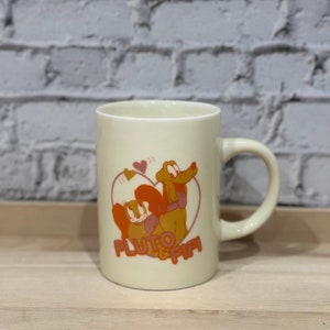 Peut inclure: Une tasse en céramique blanche avec une image de dessin animé de Pluto et Fifi dans un cœur. Le texte "Pluto & Fifi" est imprimé en orange sous l'image.