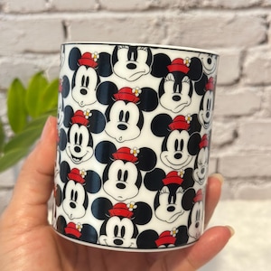 Puede incluir: Taza de cer&aacute;mica blanca con un patr&oacute;n repetido de caras de Mickey Mouse con sombreros rojos. La taza tiene un borde negro. El fondo es una pared de ladrillos blancos. La taza est&aacute; sostenida en una mano.