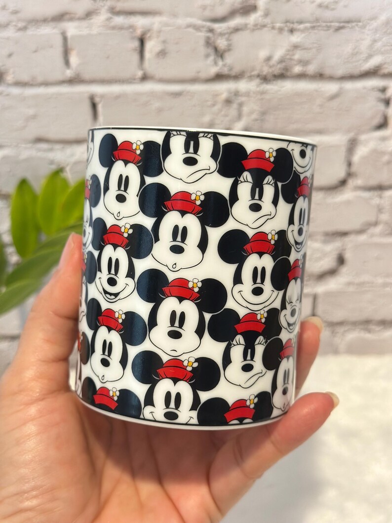 Puede incluir: Taza de cer&aacute;mica blanca con un patr&oacute;n repetido de caras de Mickey Mouse con sombreros rojos. La taza tiene un borde negro. El fondo es una pared de ladrillos blancos. La taza est&aacute; sostenida en una mano.