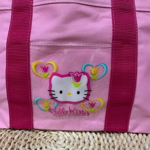 K&ouml;nnte beinhalten: Rosa Hello Kitty-Tragetasche mit rosa Henkeln und einer durchsichtigen Tasche mit einem Hello Kitty-Motiv mit Herzen und Blumen.
