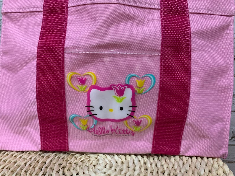 K&ouml;nnte beinhalten: Rosa Hello Kitty-Tragetasche mit rosa Henkeln und einer durchsichtigen Tasche mit einem Hello Kitty-Motiv mit Herzen und Blumen.